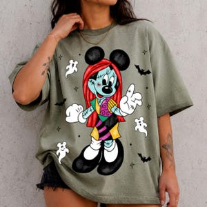 May include: An olive green short-sleeve t-shirt featuring a graphic print of a character with red hair, a patchwork dress, and black ears. The design includes white ghost figures, black bats, and small star accents surrounding the central character.