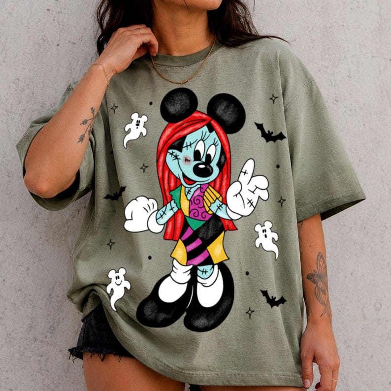 May include: An olive green short-sleeve t-shirt featuring a graphic print of a character with red hair, a patchwork dress, and black ears. The design includes white ghost figures, black bats, and small star accents surrounding the central character.