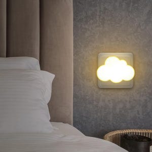 Può includere: Una luce notturna a forma di nuvola che emette una calda luce. La luce è montata su una base quadrata bianca contro un muro grigio strutturato. La scena comprende un letto con biancheria da letto bianca e un cestino di vimini.