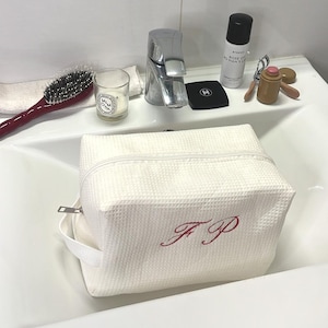 Może przedstawiać: Biała, teksturowana kosmetyczka z czerwonymi haftowanymi inicjałami "F P" leży w umywalce. Inne przedmioty to szczotka do włosów, świeca, makijaż i butelka Rose of No Man's Land.