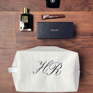 Peut inclure: Ensemble d'articles de luxe sur une surface en bois. Comprend un flacon de parfum noir, un boîtier noir avec un logo, un tube de produit pour les lèvres, une boîte Prada bleu marine et une trousse de toilette blanche avec des initiales brodées.