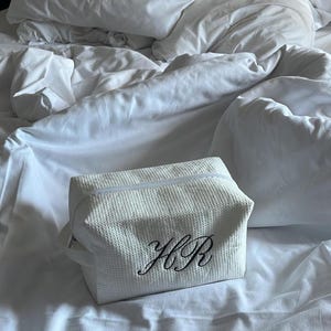 Peut inclure: Une trousse de toilette blanche à texture gaufrée dotée d'une fermeture éclair et d'une poignée latérale. L'avant de la trousse est brodé des lettres cursives noires HR. La pochette rectangulaire présente une forme structurée et un fini uniforme.