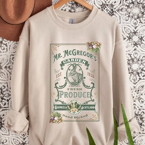 May include: Beige sweatshirt with a vintage-style graphic. The graphic reads "Mr. McGregor's Garden Fresh Produce" with a rabbit illustration. Additional text includes "Dunkeld Scotland" and "Hand Picked".