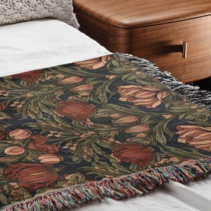 Puede incluir: Una manta de tiro negra y verde con un patrón floral y flecos. La manta está sobre una cama blanca con una almohada blanca y una mesita de noche de madera.