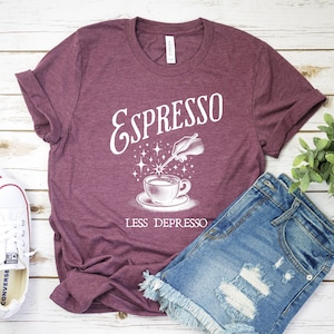 May include: A maroon t-shirt with white text that reads "Espresso Less Depresso". The text is accompanied by a graphic of a coffee cup with a magic wand hovering over it.