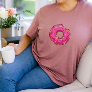 May include: A woman wearing a mauve t-shirt with a pink donut graphic. The donut has colorful sprinkles and a bite taken out of it.