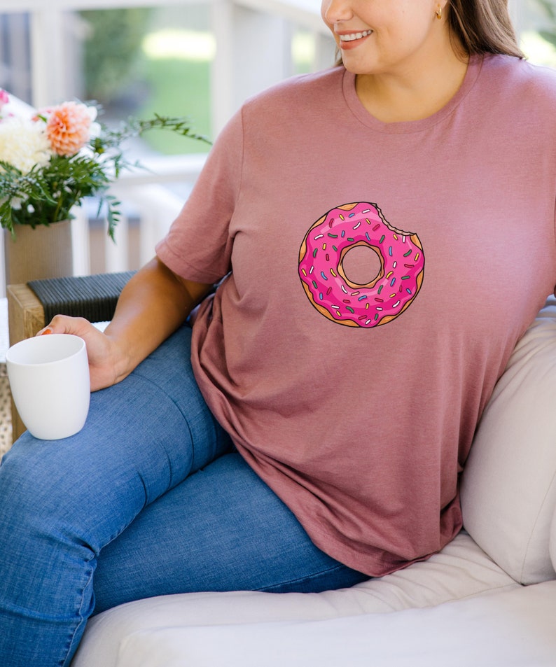 May include: A woman wearing a mauve t-shirt with a pink donut graphic. The donut has colorful sprinkles and a bite taken out of it.