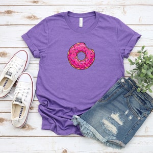 May include: Purple t-shirt with a pink donut with sprinkles and a bite taken out of it. The donut is centered on the shirt. The shirt is laying on a white wooden surface with a pair of white sneakers and a pair of denim shorts.