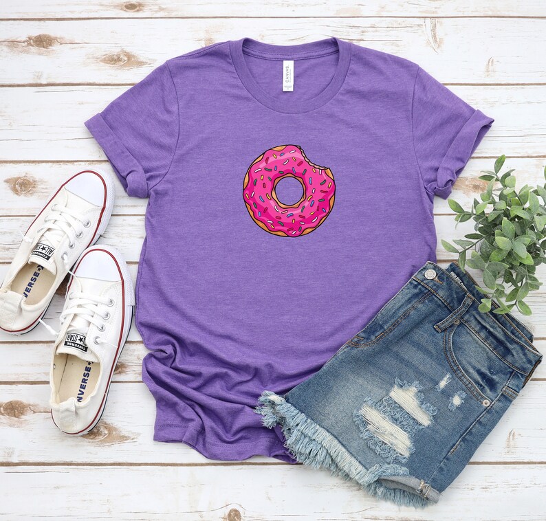 May include: Purple t-shirt with a pink donut with sprinkles and a bite taken out of it. The donut is centered on the shirt. The shirt is laying on a white wooden surface with a pair of white sneakers and a pair of denim shorts.