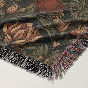 Pu&ograve; includere: Una coperta in tessuto intrecciato con uno sfondo verde scuro e un motivo floreale in tonalit&agrave; di marrone, rosso e crema. La coperta ha un bordo a frangia.