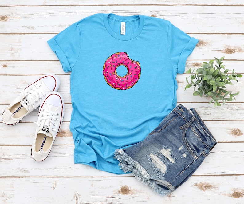 May include: A turquoise blue t-shirt with a pink and white donut graphic. The donut has sprinkles and a bite taken out of it. The t-shirt is laying on a white wooden surface with a pair of white sneakers and denim shorts.