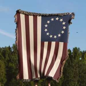 May include: A woven throw blanket featuring the design of the Betsy Ross flag. The blanket has alternating red and white stripes with a blue square in the upper left corner containing a circle of white stars. The edges are fringed.