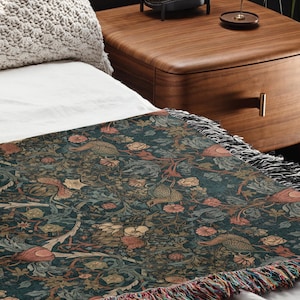 May include: A dark teal throw blanket with a detailed floral and bird pattern. The blanket has a fringed edge with multiple colors. The throw is draped over a white bed sheet.