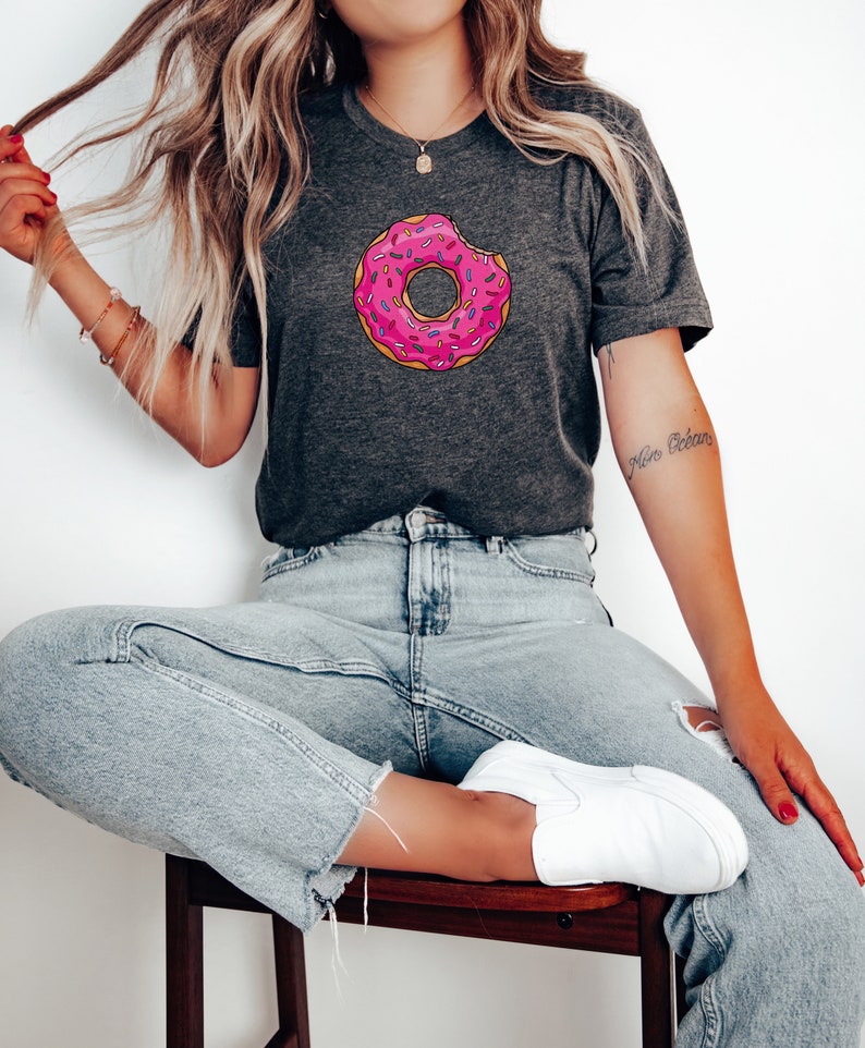 May include: A woman wearing a dark grey t-shirt with a pink and white donut graphic. The donut has sprinkles and a bite taken out of it. The woman is wearing light blue jeans and white sneakers. The text "Here We Go" is tattooed on her arm.