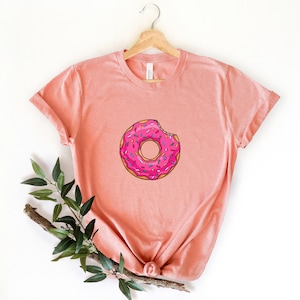 May include: A pink t-shirt with a donut graphic. The donut is pink with white frosting and colorful sprinkles. A bite has been taken out of the donut.