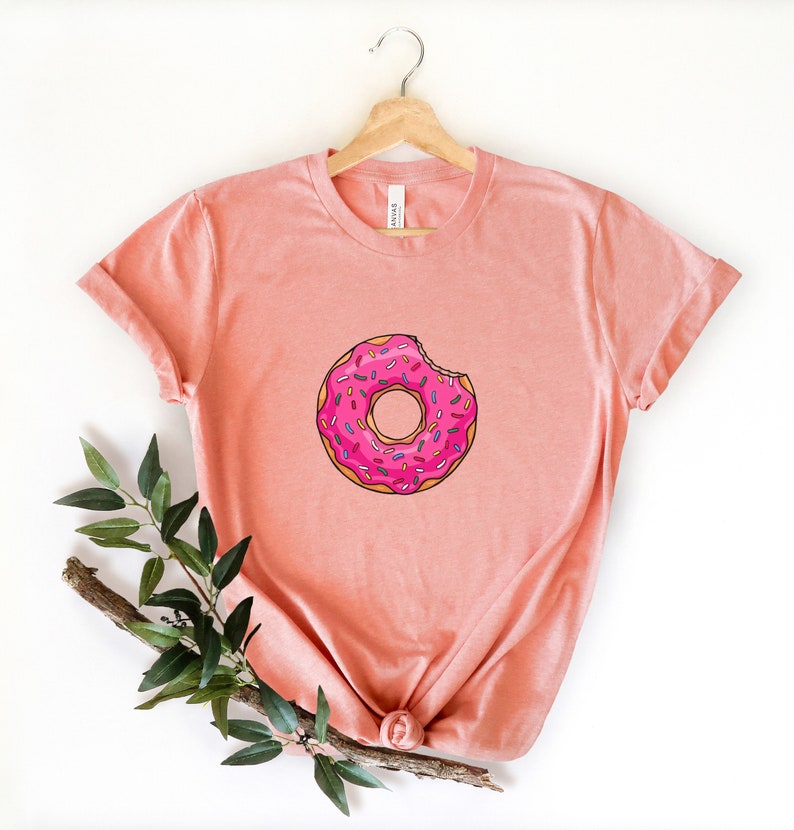 May include: A pink t-shirt with a donut graphic. The donut is pink with white frosting and colorful sprinkles. A bite has been taken out of the donut.