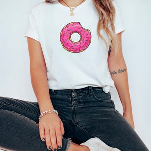 May include: A white t-shirt with a pink and white donut graphic. The donut has sprinkles and a bite taken out of it.