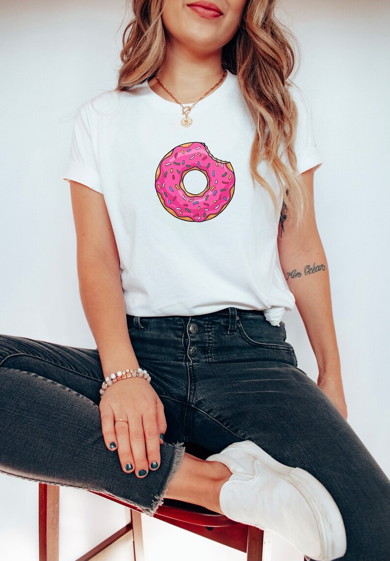 May include: A white t-shirt with a pink and white donut graphic. The donut has sprinkles and a bite taken out of it.
