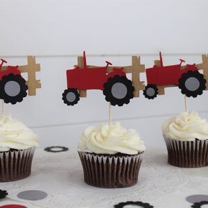 K&ouml;nnte beinhalten: Drei Cupcakes mit wei&szlig;em Zuckerguss, jeweils gekr&ouml;nt von einem roten Traktor und einem braunen Zaun. Die Traktoraufs&auml;tze haben schwarze und graue R&auml;der. Konfetti in Schwarz, Grau und Rot umgibt die Cupcakes.