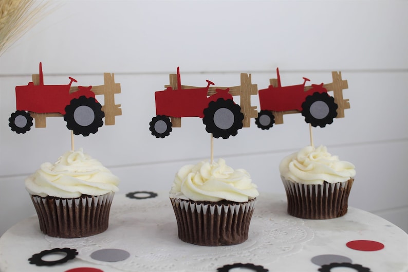 K&ouml;nnte beinhalten: Drei Cupcakes mit wei&szlig;em Zuckerguss, jeweils gekr&ouml;nt von einem roten Traktor und einem braunen Zaun. Die Traktoraufs&auml;tze haben schwarze und graue R&auml;der. Konfetti in Schwarz, Grau und Rot umgibt die Cupcakes.