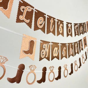 May include: A banner with brown and gold glittery pennants strung on white string. The banner says "Let's Get Nashed" with images of cowboy boots and diamond rings. The banner is for a bachelorette party.