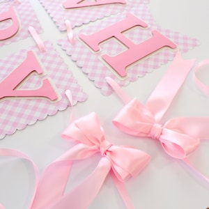 May include: Pink banner with scalloped edges and a gingham pattern. The banner features pink letters and is adorned with pink satin bows. The letters are attached to the banner with pink ribbons.