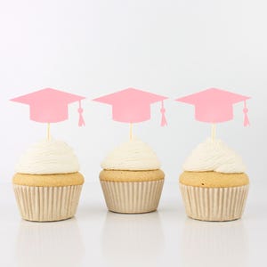 May include: Three cupcakes with white frosting and pink graduation cap toppers. The cupcakes have a light brown base and are in a fluted paper liner. The graduation caps are pink with a tassel.