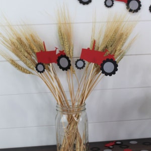 May include: Red and gray paper tractor cutouts on sticks in a glass jar with wheat stalks. The tractor cutouts are scattered on a wooden table with red and gray circles.