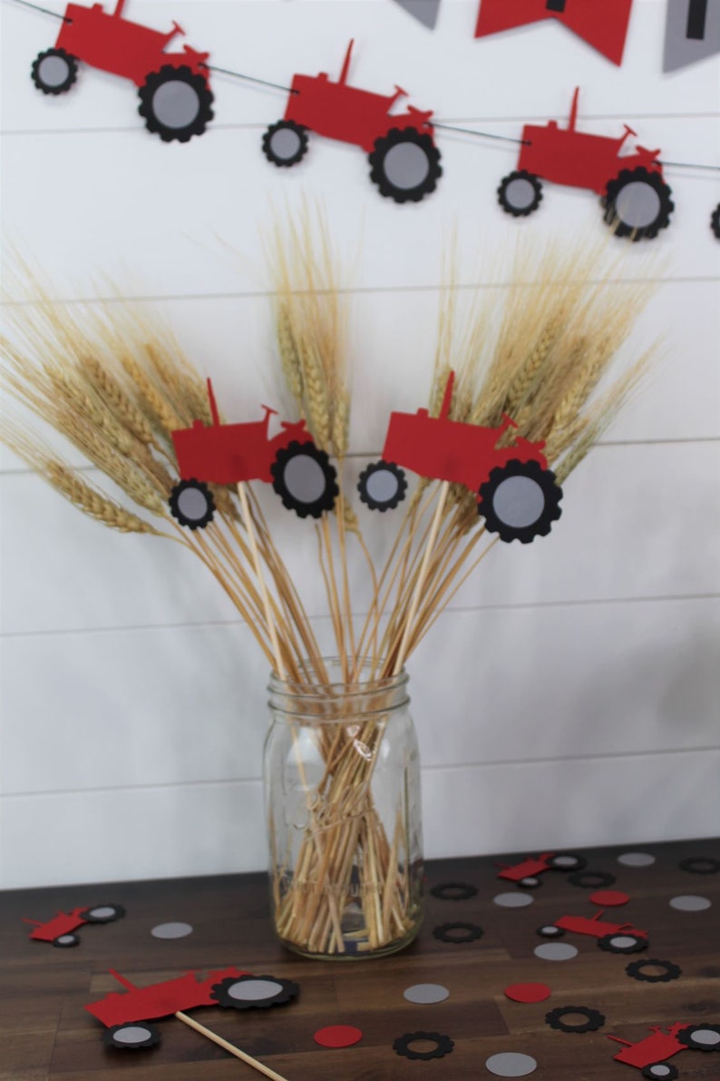 May include: Red and gray paper tractor cutouts on sticks in a glass jar with wheat stalks. The tractor cutouts are scattered on a wooden table with red and gray circles.
