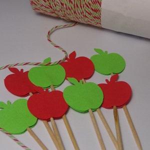 May include: Red and green paper apple cutouts on wooden skewers. The apples are arranged in a semi-circle on a white surface.