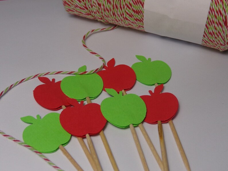 May include: Red and green paper apple cutouts on wooden skewers. The apples are arranged in a semi-circle on a white surface.
