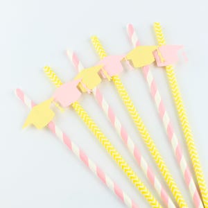 May include: A collection of graduation-themed paper straws. The straws feature pink and white stripes, and yellow and white chevron patterns. Each straw is topped with a pink or yellow graduation cap cutout.