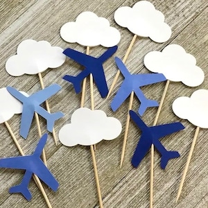 May include: Blue and white paper airplane and cloud cake toppers. The airplanes are blue with white outlines and the clouds are white with a slight blue tint. The toppers are attached to wooden sticks.