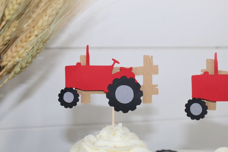 May include: Red and black tractor cupcake topper with a brown wooden fence. The tractor has a gray wheel and a black tire.
