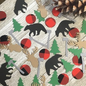 May include: A collection of rustic-themed paper cutouts, including black bears, moose, green trees, and gray axes, scattered on a light wood-grain surface. Red and black plaid circles add a pop of color. A pine cone rests in the corner.