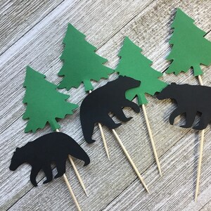 May include: Set of six cupcake toppers featuring black bear silhouettes and green paper evergreen trees. Each topper is attached to a wooden skewer. Ideal for a woodland or camping-themed party.