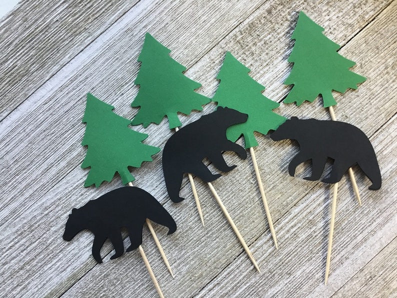 May include: Set of six cupcake toppers featuring black bear silhouettes and green paper evergreen trees. Each topper is attached to a wooden skewer. Ideal for a woodland or camping-themed party.