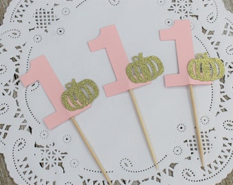 Peut inclure: Trois toppers de cupcake roses avec le chiffre "1" et des citrouilles dorées à paillettes.