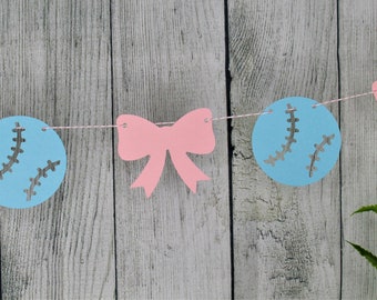 May include: A blue and pink gender reveal banner with baseballs and bows. The baseballs have white stitching and the bows are pink.