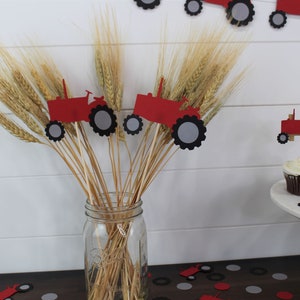 K&ouml;nnte beinhalten: Ein klares Glasgef&auml;&szlig; gef&uuml;llt mit Weizen&auml;hren und roten Traktordekorationen. Die Traktor-Ausschnitte haben schwarze und graue R&auml;der. Cupcakes mit wei&szlig;em Zuckerguss und Traktor-Toppern stehen auf einem wei&szlig;en Kuchenst&auml;nder.