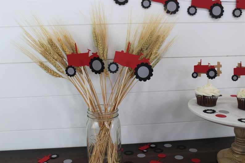K&ouml;nnte beinhalten: Ein klares Glasgef&auml;&szlig; gef&uuml;llt mit Weizen&auml;hren und roten Traktordekorationen. Die Traktor-Ausschnitte haben schwarze und graue R&auml;der. Cupcakes mit wei&szlig;em Zuckerguss und Traktor-Toppern stehen auf einem wei&szlig;en Kuchenst&auml;nder.