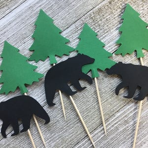May include: A collection of paper cutouts featuring black bear silhouettes and green evergreen trees attached to wooden skewers. These decorative elements are likely intended for use as cake toppers or party decorations, evoking a woodland theme.