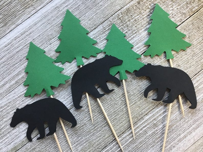 May include: A collection of paper cutouts featuring black bear silhouettes and green evergreen trees attached to wooden skewers. These decorative elements are likely intended for use as cake toppers or party decorations, evoking a woodland theme.