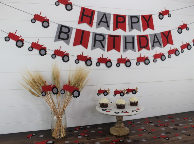 May include: Red and gray tractor themed birthday party decorations. A banner reads "Happy Birthday" with red and gray pennants and red tractors. There are also red tractor cupcake toppers and red and gray confetti.