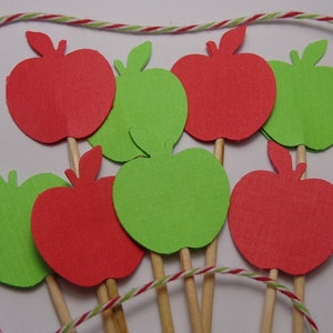 May include: A set of red and green apple-shaped paper cutouts on wooden sticks. The apples are arranged in a circle and are ready to be used as cake toppers or party decorations.