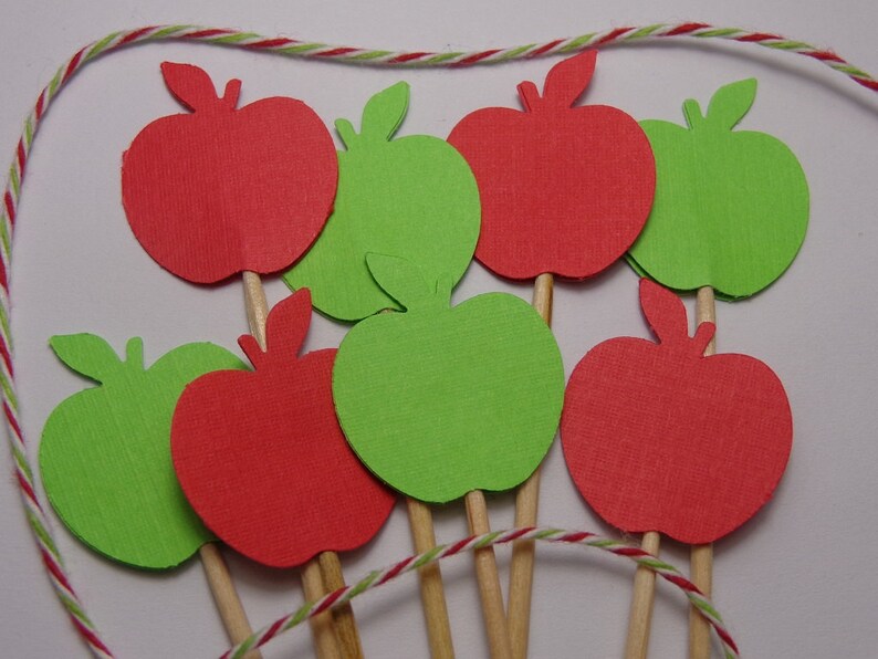 May include: A set of red and green apple-shaped paper cutouts on wooden sticks. The apples are arranged in a circle and are ready to be used as cake toppers or party decorations.