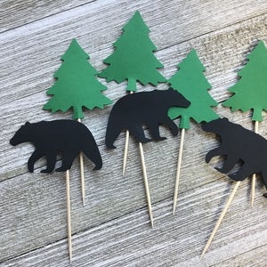 May include: Set of cupcake toppers featuring black bear silhouettes and green paper evergreen trees. Each topper is attached to a wooden skewer. The toppers are arranged on a light wood grain surface.