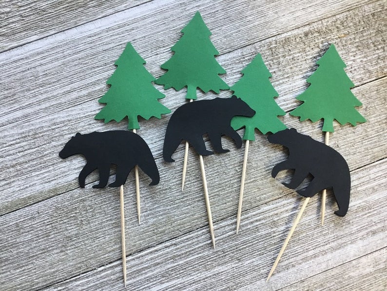 May include: Set of cupcake toppers featuring black bear silhouettes and green paper evergreen trees. Each topper is attached to a wooden skewer. The toppers are arranged on a light wood grain surface.