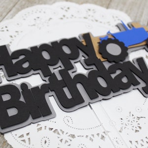 May include: Black and gray paper cake topper with the words "happy birthday" and a blue tractor with a gray tire.