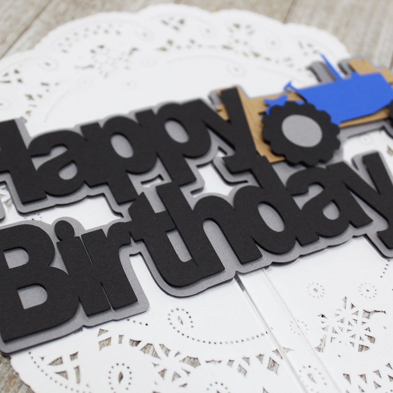 May include: Black and gray paper cake topper with the words "happy birthday" and a blue tractor with a gray tire.